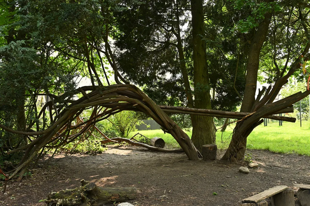Hof der Kinderkreisel-Kita Waldgeistergruppe mit bogenförmigem Baum und verbundenen Baumstämmen zum Balancieren.