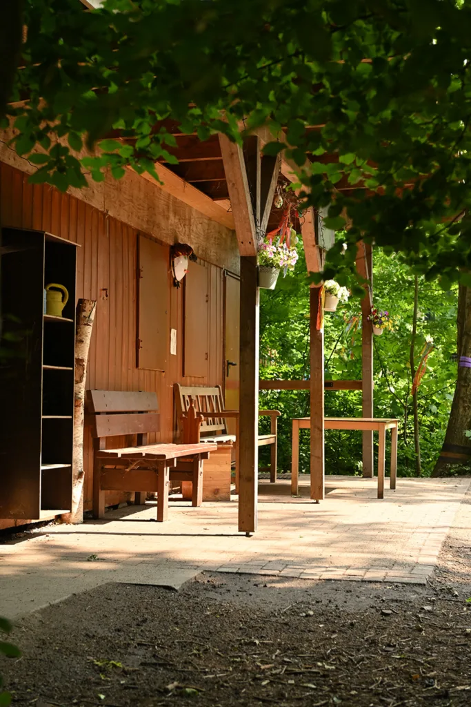 Rotes Holz-Außengebäude der Kinderkreisel-Kita Waldgeistergruppe mit Bänken, Tisch und Regal für Gießkannen.