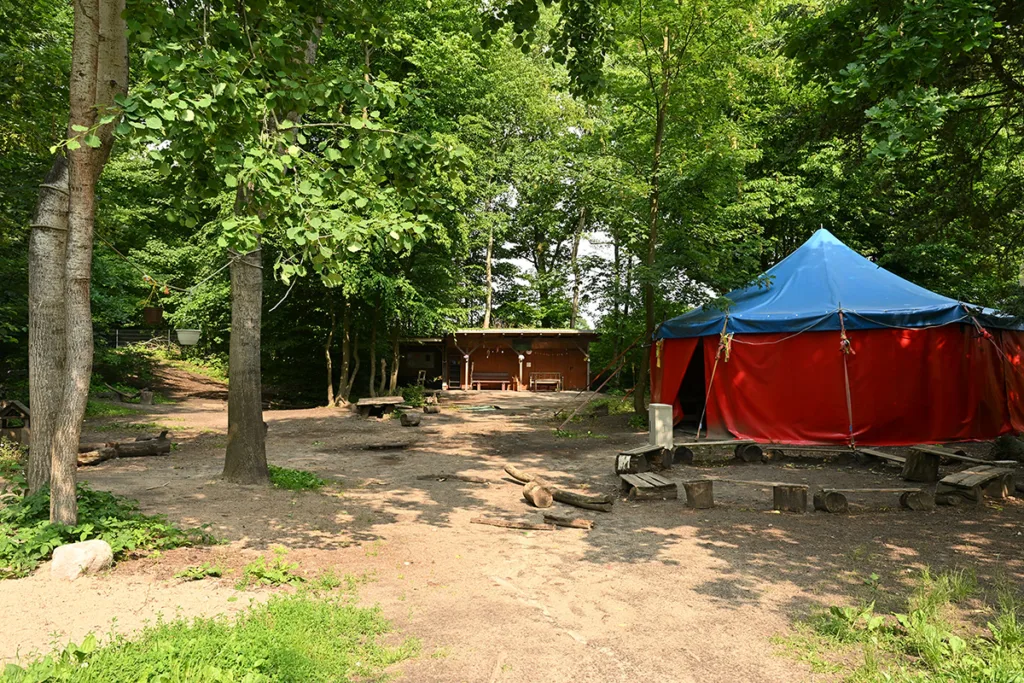 Hof der Kinderkreisel-Kita Waldgeistergruppe mit großem Zelt, Blick auf das rote Holzgebäude und einem Kreis aus Holzbänken davor.