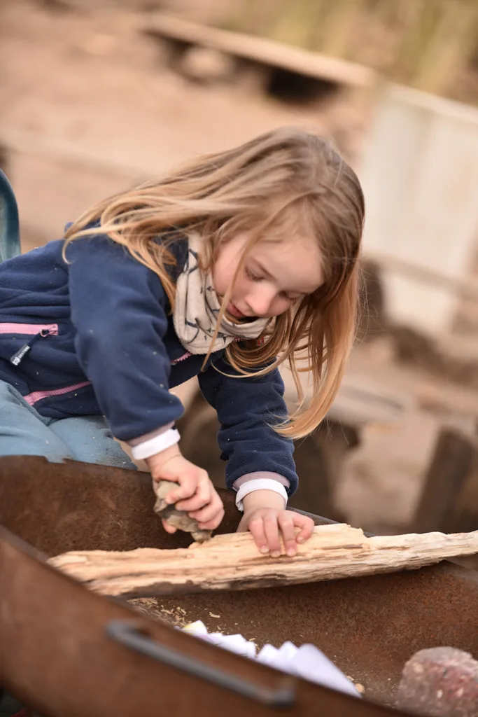 Kind legt Feuerholz in eine Feuerschale und beteiligt sich an einer begleiteten Aktivität im Außenbereich.