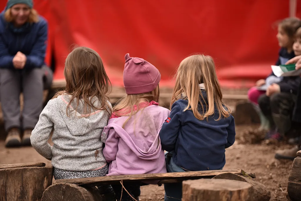 Kinder sitzen mit einer Erzieherin im Sitzkreis auf Holzbänken und erleben gemeinschaftliche Zeit.
