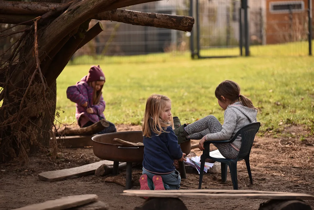 Drei Kinder sitzen um eine Feuerschale herum und kümmern sich gemeinsam um das Feuerholz.