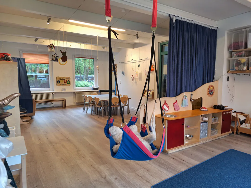 Gruppenraum der Kinderkreisel-Kita Pinguinegruppe mit Hängematte, Regalen zur Aufbewahrung von Spielzeug und Materialien sowie Tischen im Hintergrund.