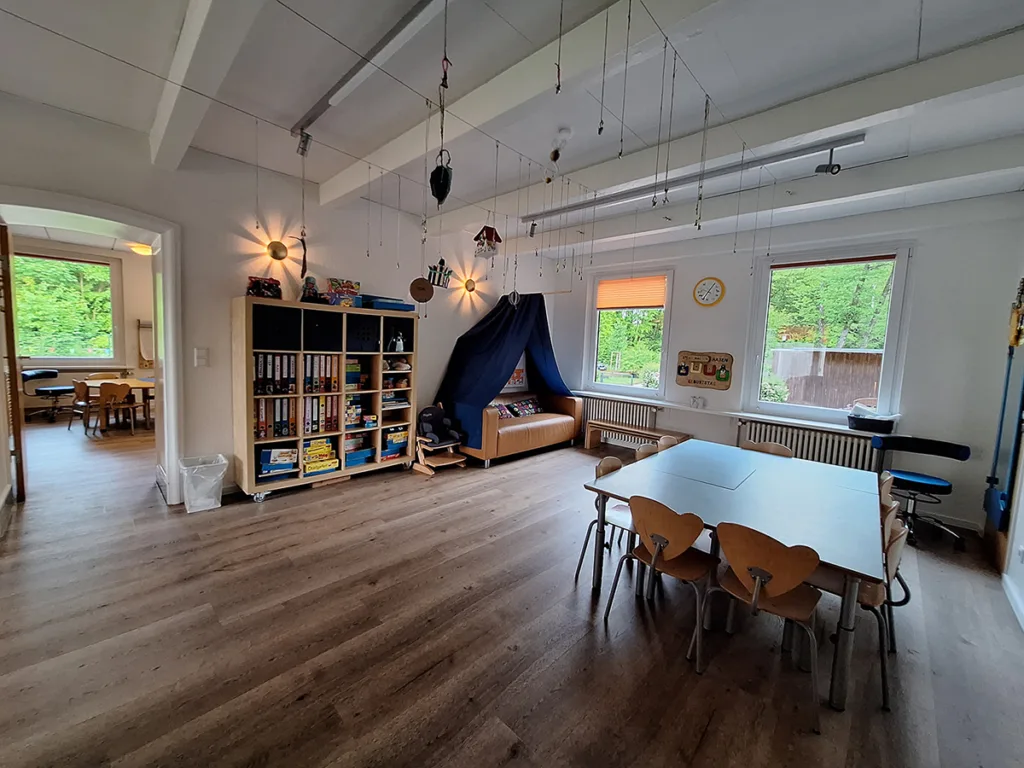 Gruppenraum der Kinderkreisel-Kita Pinguinegruppe mit großem Tisch und vielen Stühlen, Sofa mit zuziehbarer Gardine sowie großem Regal mit Ordnern und Brettspielen.