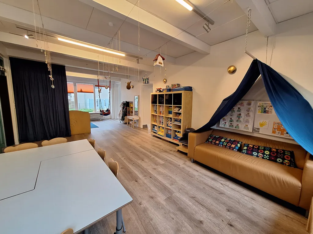 Gruppenraum der Kinderkreisel-Kita Pinguinegruppe mit großem Tisch und vielen Stühlen, Sofa mit zuziehbarer Gardine sowie großem Regal mit Ordnern und Brettspielen.