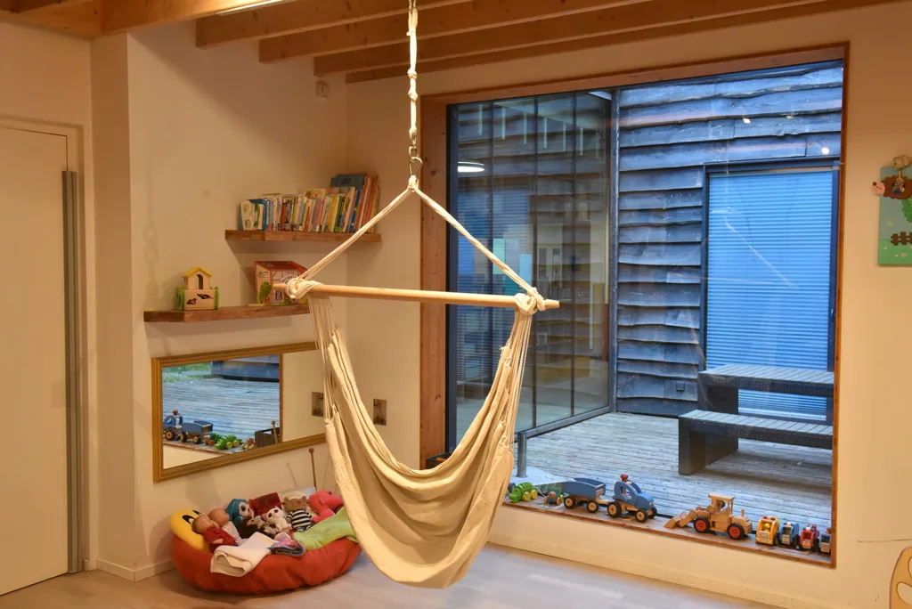 Gemütliche Ecke in der Kinderkreisel-Kita Igelgruppe mit Hängematte, Kinderbüchern und Spielzeug am großen Fenster mit Blick in den Innenhof mit Gartentisch und Bänken.