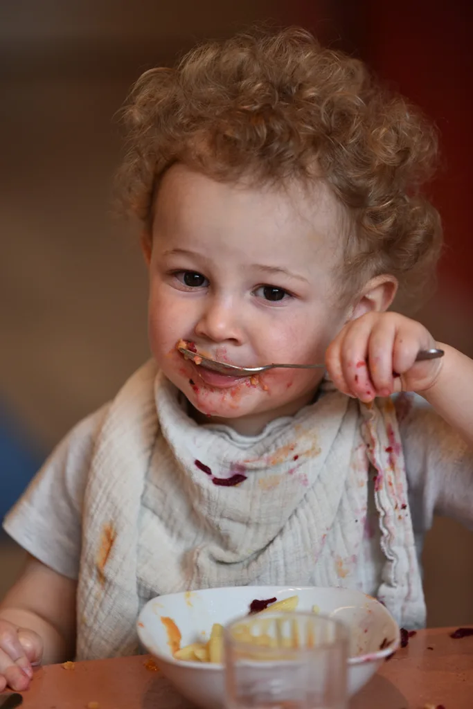 Kleinkind isst selbstständig ein Nudelgericht mit dem Löffel und trägt ein mit Essen bekleckertes Tuch um den Hals.