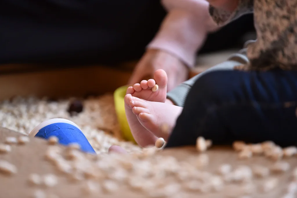 Kleinkind sitzt auf dem Boden und erkundet spielerisch kleine Kerne, die auf dem Boden verteilt und zwischen den Zehen liegen.