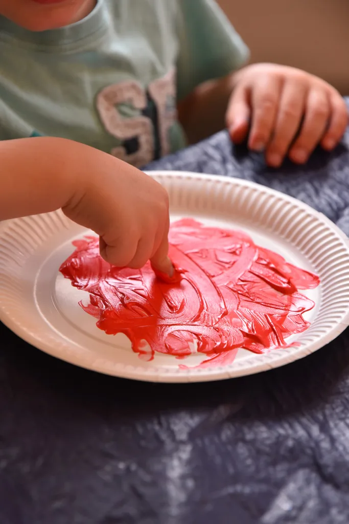 Kleinkind experimentiert mit roter Fingerfarbe und malt mit den Fingern auf einen Pappteller.