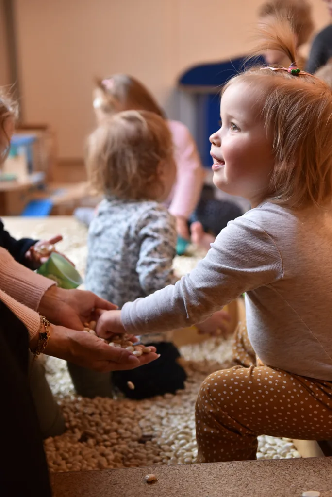 Kleinkind nimmt freudig kleine Kerne aus der Hand der Erzieherin und sammelt dabei sensorische Erfahrungen, während weitere Kerne auf dem Boden liegen.