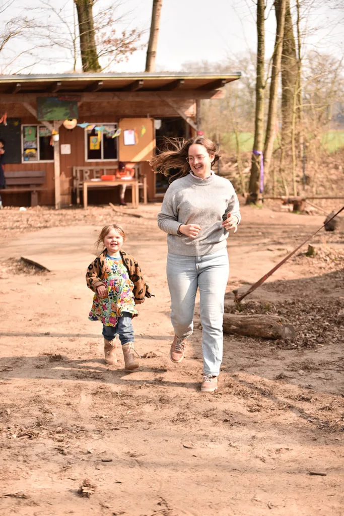 Erzieherin läuft gemeinsam mit einem Kind über Erdboden, im Hintergrund eine Hütte mit Bänken.